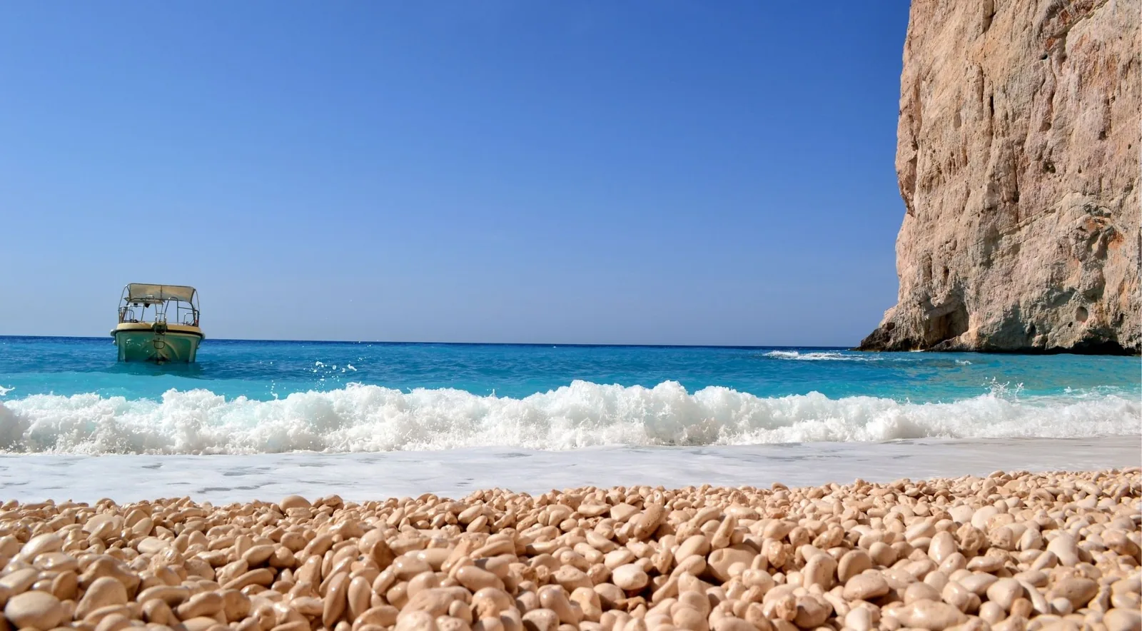 The shore at Navagio in close-up: rounded white limestone pebbles, with a small boat offshore.