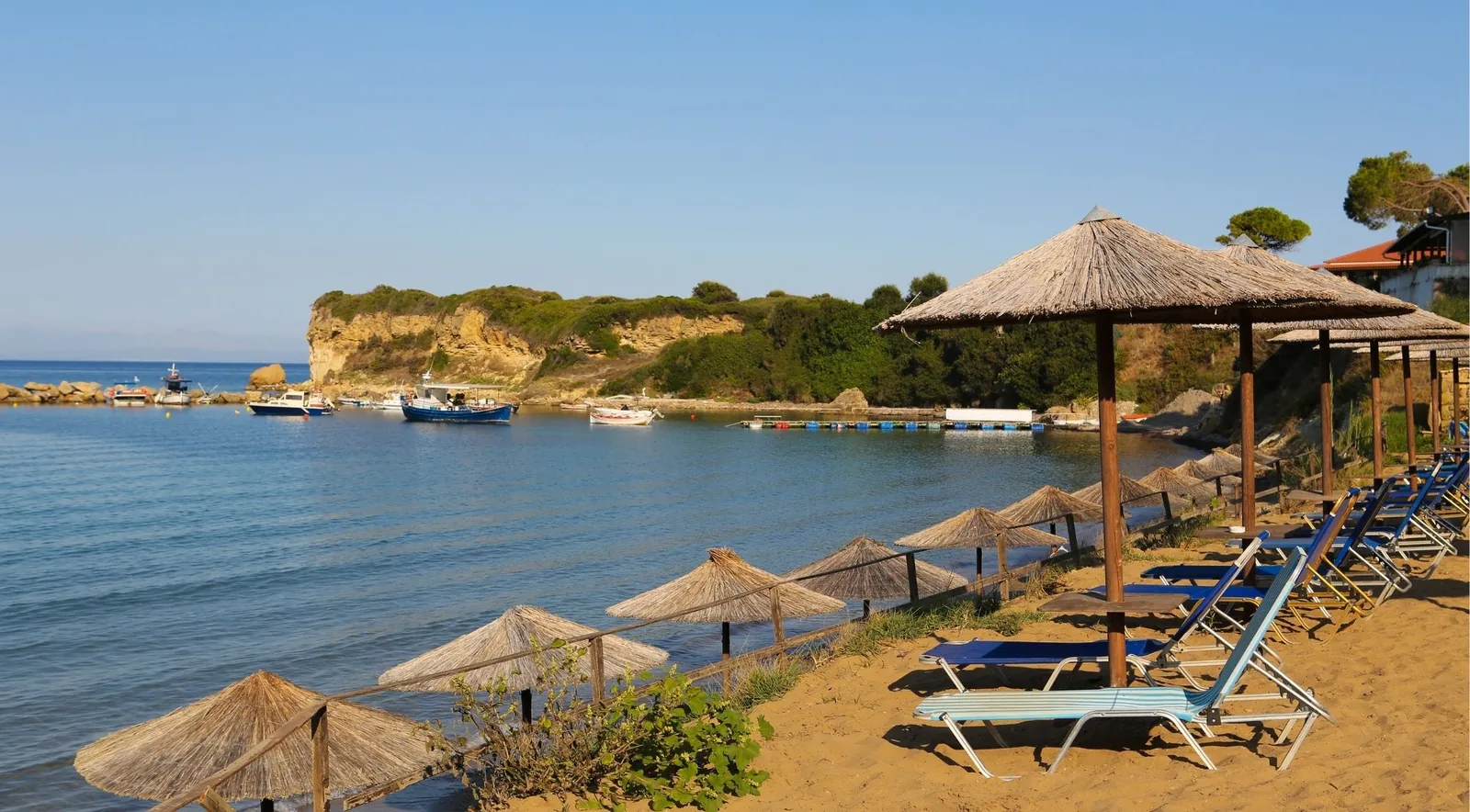 A small sandy beach with thatched umbrellas beside a fishing harbour, a low headland behind.