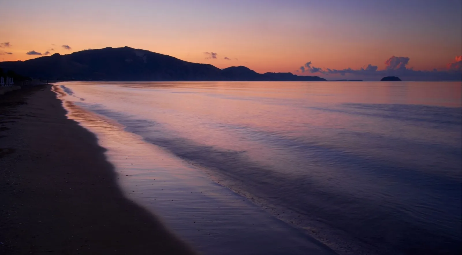 Laganas Bay at dusk, the wet sand reflecting orange light, with a low island silhouetted offshore.
