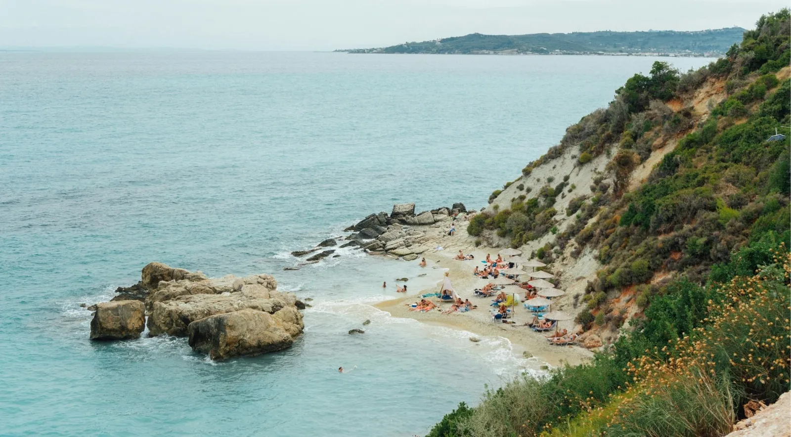 A small cove of pale pebbles below scrubby cliffs, umbrellas set out along the shoreline.