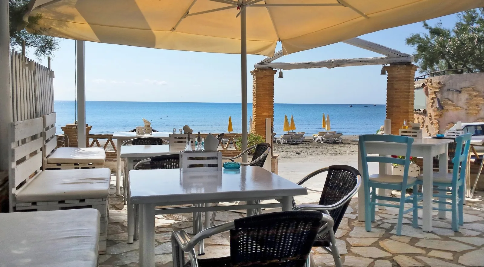 A taverna terrace with white tables under an awning, looking out over sand and flat sea.
